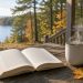 Open book and steaming mug on a wooden porch table with autumn maple leaves and a lake beyond