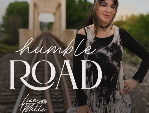 Woman with long dark hair standing beside railroad tracks that lead toward a metal truss bridge, wearing a black-and-white fringed tunic in warm evening light