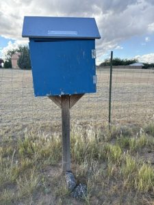 A small blue Little Free Library box on a wooden post beside a wire fence and a dry grassy field under a cloudy sky in Chino Valley.