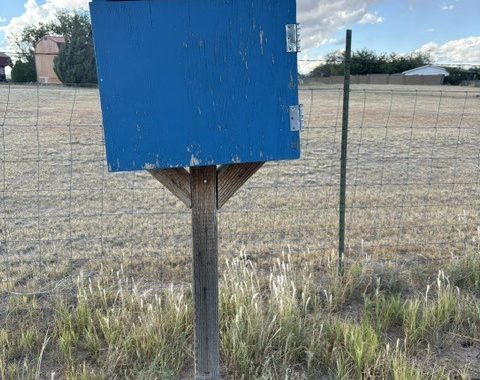 A small blue Little Free Library box on a wooden post beside a wire fence and a dry grassy field under a cloudy sky in Chino Valley.