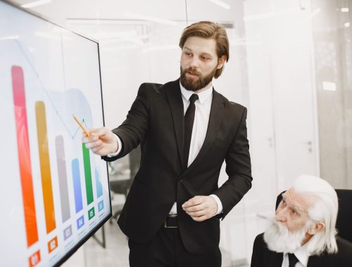 Financial advisor in a suit pointing to a bar chart on a screen while a seated older businessman reviews the numbers.