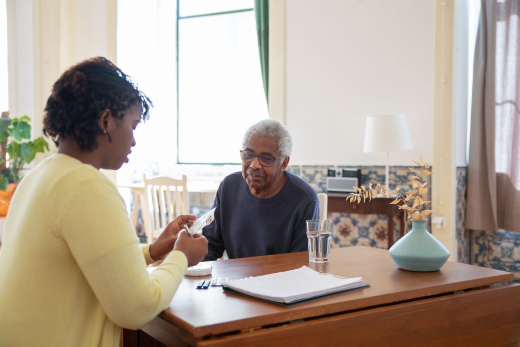 Caregiver in a yellow sweater sorting pills into a pill organizer for an older man at a dining table.