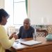 Caregiver in a yellow sweater sorting pills into a pill organizer for an older man at a dining table.