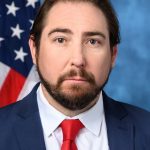 Official portrait of U.S. Rep. Eli Crane of Arizona in a navy suit and red tie in front of an American flag.